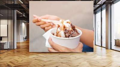 Outdoor street closeup view of hand hold a cup, plastic spoon, and eat melting frozen yogurt ice cream with chocolate topping in white paper cup. Wall mural