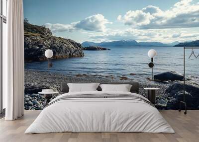 A calm, pebbled beach with a clear blue sky, rocky shores, and a view of mountains in the distance. Wall mural