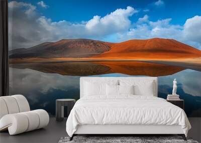 A clear blue sky and white clouds are reflected in a calm lake, surrounded by red volcanic hills and a sandy beach. Wall mural