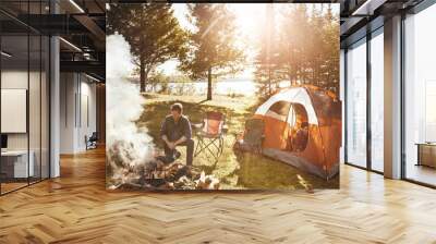 Couple, campfire and wood with smoke in nature by tent on grass field for outdoor vacation, holiday or weekend. Man preparing fire by woman for camping leisure, summer getaway or sunshine at forest Wall mural