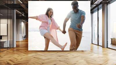 Laugh, holding hands and splash with interracial couple at beach for travel destination, summer or honeymoon vacation. Relax, tropical and adventure with man and woman for holiday, bonding or romance Wall mural