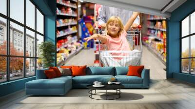 Little girl having fun riding in a shopping cart while shopping groceries at the supermarket with her mother Wall mural