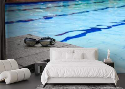 A pair of swimming goggles rests on the edge of a swimming pool, with the clear blue water in the background. Swimming Goggles by the Pool Background. Wall mural