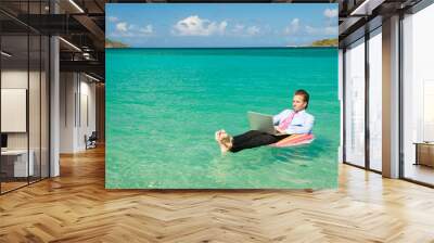 Barefoot businessman working on his laptop while floating on a colorful inflatable ring in bright tropical waters Wall mural