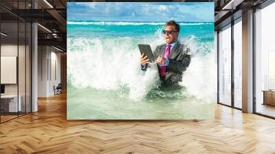 Fearful businessman using his tablet computer with oncoming waves crashing on the shore of a tropical beach   Wall mural