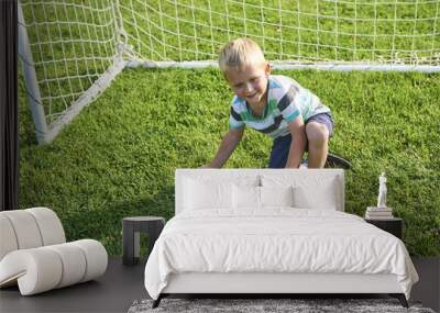 Cute little blond boy playing at being a goalkeeper on a sportsfield standing in the goalposts in sunny summer day

 Wall mural