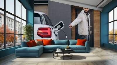 Man charging electric car by the house Wall mural