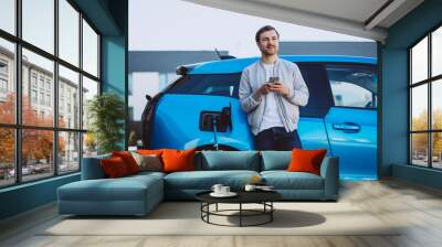 Young man charging electric car at home and using mobile phone Wall mural