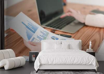 A person analyzes financial graphs and charts while working on a laptop on a wooden table, indicating a focus on data evaluation and reporting. Wall mural