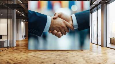 Two businessmen confidently shaking hands in front of a blurred office backdrop adorned with vibrant flags, symbolizing trust, agreement, and successful global collaboration. Wall mural