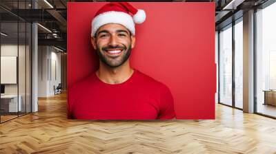 A cheerful man dressed in a red shirt and Christmas hat stands against a vibrant red wall, embodying holiday spirit Wall mural