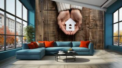 A close-up of two hands holding a paper house cutout on a rustic wood background Wall mural