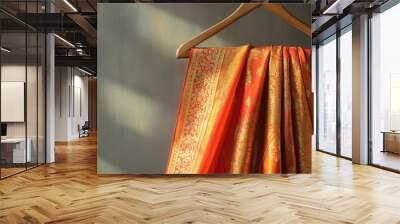 A vibrant orange silk sari with intricate zari embroidery, gracefully flowing on a wooden hanger, plain gray background. Wall mural