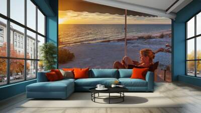 Retired couple sitting under the awning of a of caravan enjoying a wine next the beach with a golden sunset. Wall mural