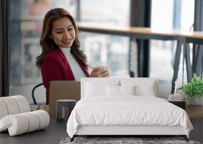 Beautiful asian businesswoman sitting working with laptop computer on table, holding cup of coffee, taking break, relaxing after working on finances. and marketing planning in the office. Wall mural