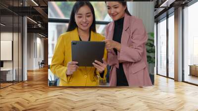 Two confident Asian businesswomen stand holding tablets to discuss working on investment projects and strategic planning together at the office. brainstorming ideas. Wall mural