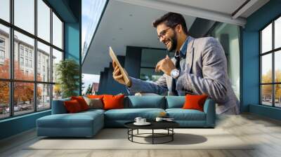 Happy young adult man in suit in front of office looking happily at digital tablet  Wall mural