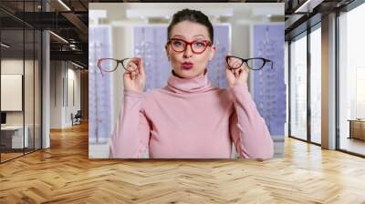 Young adult girl, choosing between two glasses in optic store and making cute face  Wall mural