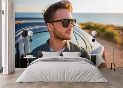 Close up of young male traveler wearing sunglasses standing next to his car on the seaside beach Wall mural