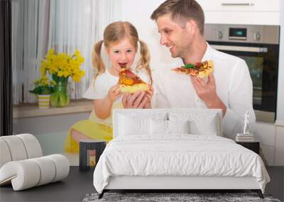 Cute little girl and her handsome father are eating pizza and smiling in the kitchen at home. Wall mural