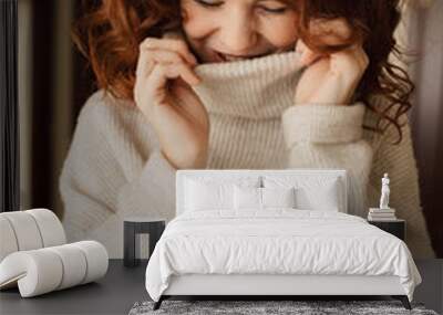 Charming cute lady with curls wearing knitted pullover is laughing and looking down with closed eyes. Indoor portrait of romantic pretty lady Wall mural