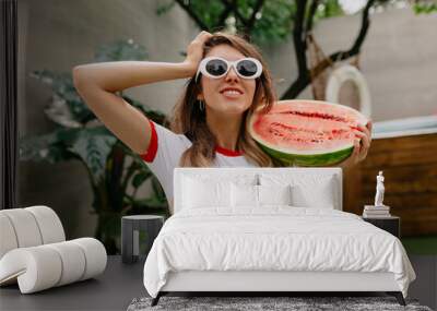 Close up portrait of lovely cute woman with wonderful smile is wearing white top and glasses is holding watermelon and touching her hair on background of exotic resort  Wall mural