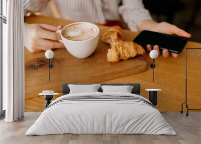 Flat lay of cappuccino with croissant on wood table. Woman hand is holding cup of coffee and smartphone  Wall mural