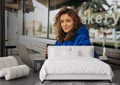 Lively young caucasian girl drinking coffee in morning and using her laptop. Brunette woman in casual clothes sits on summer terrace. Free time concept Wall mural