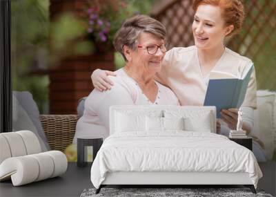 cheerful senior woman with her tender caretaker reading a book together while relaxing outside. clos Wall mural