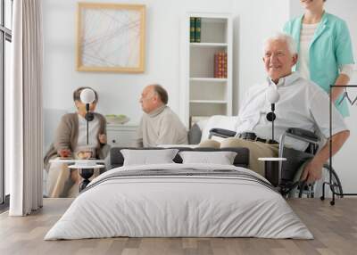 Friendly nurse in blue uniform supporting disabled elderly man on wheelchair in the nursing house Wall mural