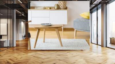 Grey armchair with patterned cushion in the real photo of bright sitting room interior with wooden small table standing on carpet, white cupboard with decor and yellow pillow on pouf Wall mural