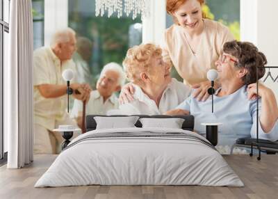 Happy friendship in old age. Tender caregiver standing behind senior women hugging each other in a nursing home. Men in the blurred background. Wall mural