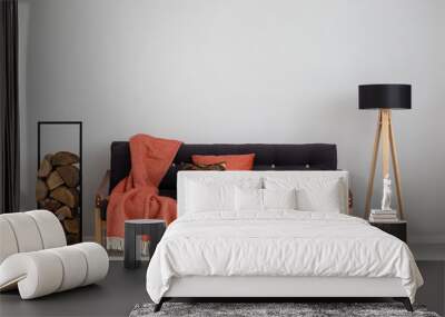 Log of wood next to comfortable sofa with strawberry red blanket and pillows, stylish wooden lamp with black lampshade, real photo copy space on the empty grey wall Wall mural