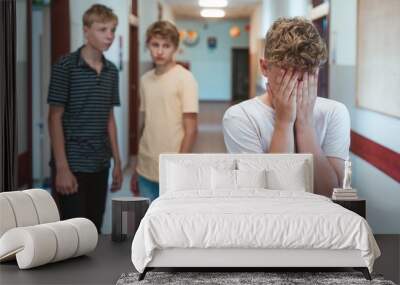 Sad bullied boy standing n the school corridor, covering his face. Two classmates behind him Wall mural