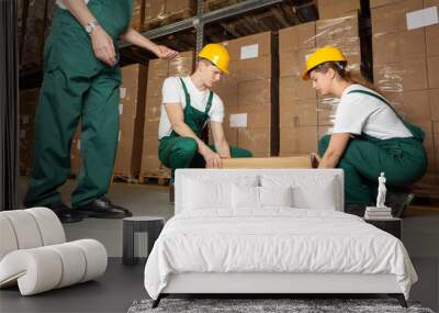 Two young warehouse workers in dark green uniforms and yellow helmets lifting heavy package Wall mural