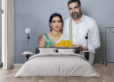 Indian couple in ethnic wear with full of sweets plate in hand looking towards the camera, south indian couple with plate of sweets in hands festival celebration Wall mural