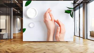 Cosmetic cream on female hands, jars with milk swirl cream and green leaves on white marble table. Flat lay, top view. Woman applying organic moisturizing hand cream. Hand skin care concept Wall mural