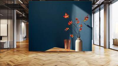 Minimalist home decor with vibrant orange flowers in a white vase, orange glass, and blank paper cad in plate, set against a deep blue wall Wall mural