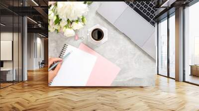 Stylish concrete stone office desk table with laptop computer, cup of coffee, flowers, female hand writing text in paper diary notepad. Top view with copy space, flat lay. Feminine workspace concept Wall mural