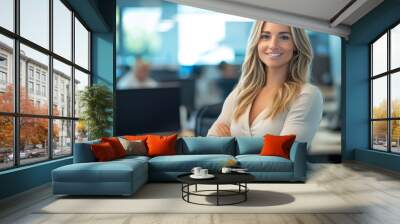 A businesswoman with blonde long hair stands in a modern office setting, with colleagues working in the background. The image captures a professional environment and leadership concept Wall mural