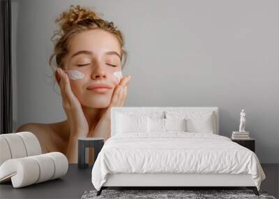 Portrait photo of a beautiful woman applying moisturizer cream on her face. Skin care morning routine. High-quality photo of younger girl on grey background. Wellness concept. Wall mural