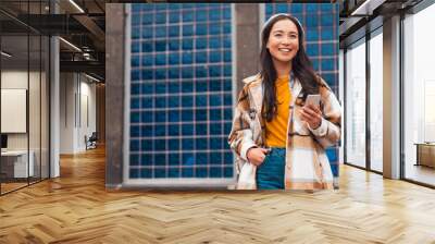 A beautiful young Asian woman is holding her phone and smiles in front of a blue tiled wall, wearing yellow t-shirt, jeans and beret.  Wall mural