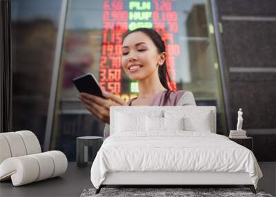 A young beautiful asian woman using an application in her smart phone to check currency exchange rates in front of an illuminated information board. Communicate about money value, international Wall mural