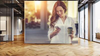 beautiful young woman is using an app in her smartphone device to send a text message in front of a sunset background Wall mural