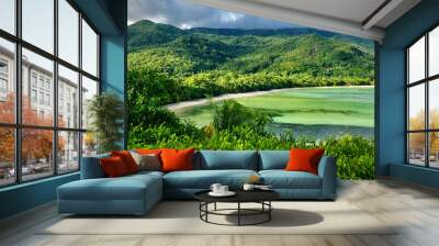 Green hills and a serene beach with clear water in Seychelles, under a cloudy sky Wall mural