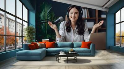 A beautiful young woman with headphones on, smiling and sitting in front of the mic for podcasting at home Wall mural