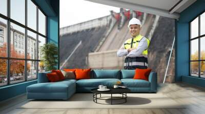 Smiling young Asian civil engineer standing with his arms crossed wearing a helmet and using a walkie-talkie. View and inspect the operation of the dam's sluice gates using the Quality Monitor tablet. Wall mural