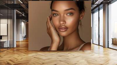 A beautiful woman with her hand touching the side of her face, wearing a light beige sleeveless top, posing for a beauty commercial in a studio setting against a brown background Wall mural