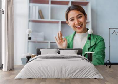 Looking to camera, Successful young pretty Asian woman waving hello talking on a video call when using laptop computer at desk in office workplace. Wall mural