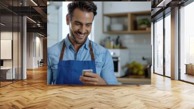 A handsome smiling man holding a cell phone in his hand standing in his kitchen Wall mural